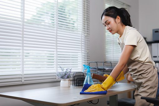 Young asian woman wearing gloves is diligently cleaning a desk in a modern office with blinds, ensuring a hygienic workspace